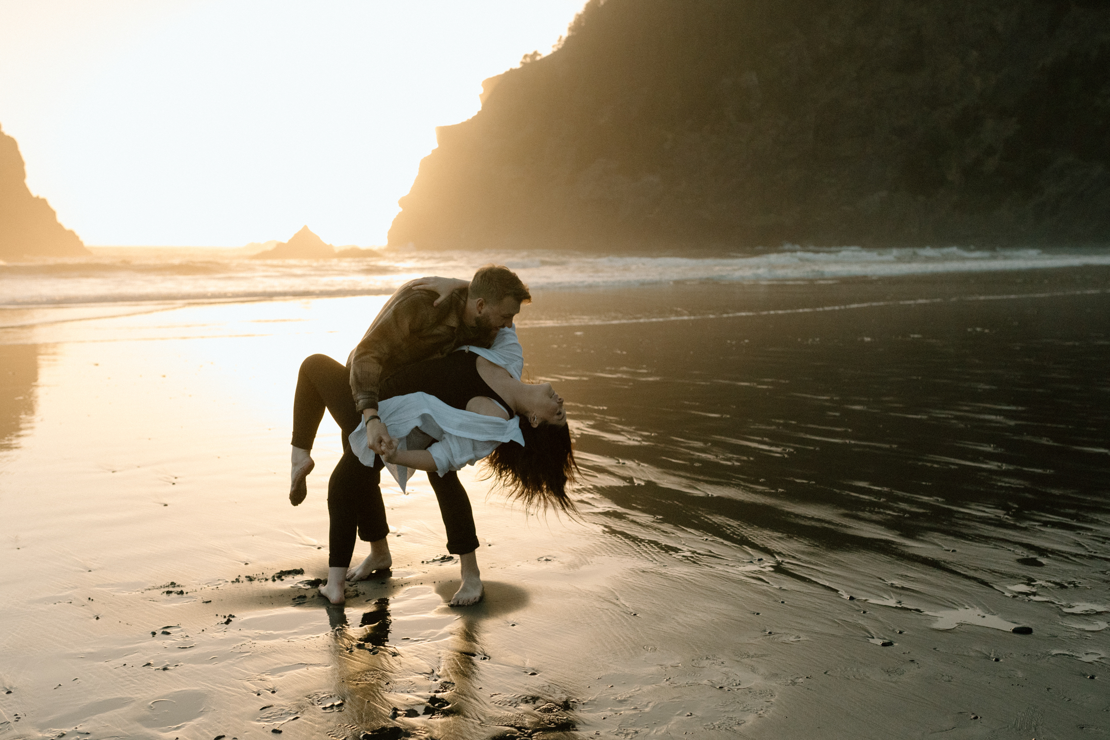 man dipping his fiance back as she kicks her leg up at whaleshead beach, oregon