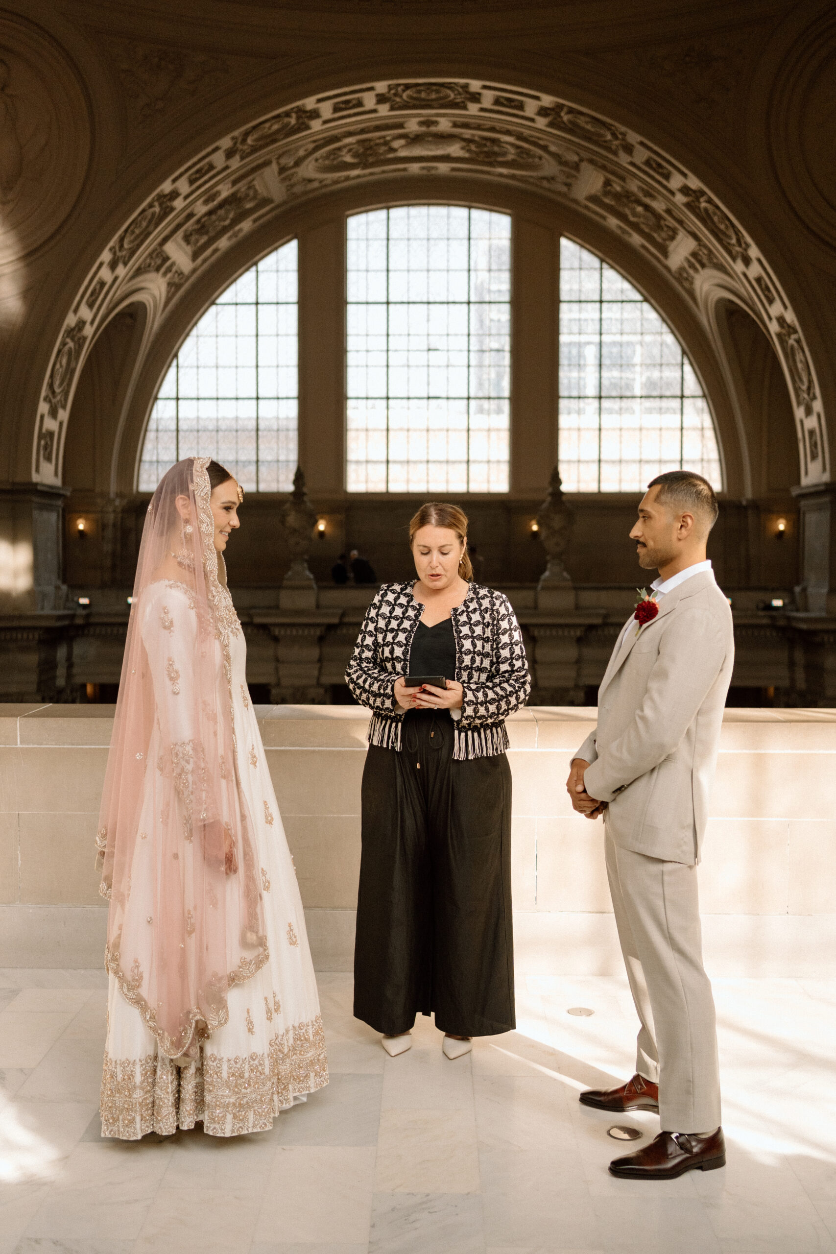 A woman in a white dress with gold embroidery and a pink headress standing in front of and smiling at a man in a tan suit with a wedding officiantin the background 