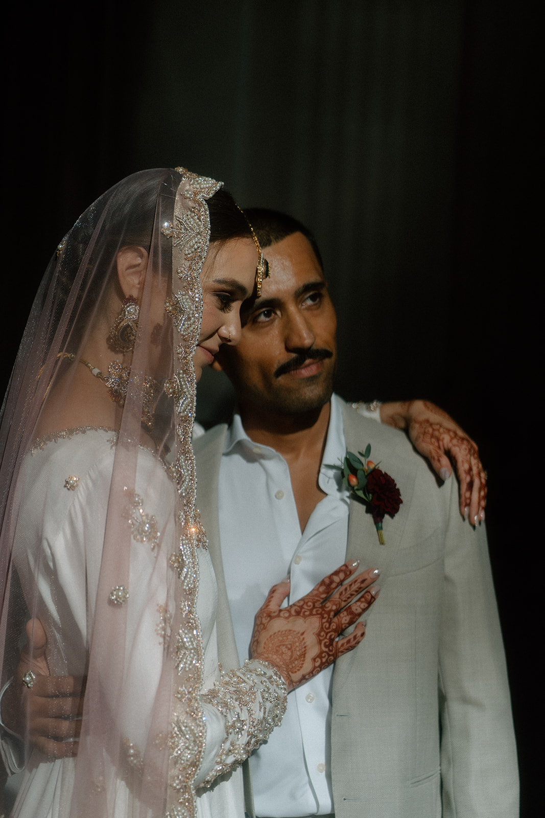 A woman with a pink headress, henna, earrings, and a white dress is touching her forehead to her partner's who is wearing a tan suit with a white button up and a boutonniere