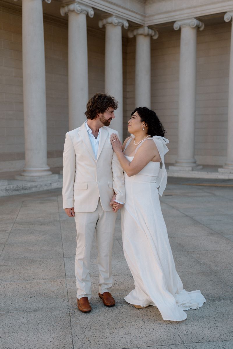 A woman in a white wedding dress is touching the shoulder of a man in a tan tuxedo they are both looking at each other behind them are tall marble columns 