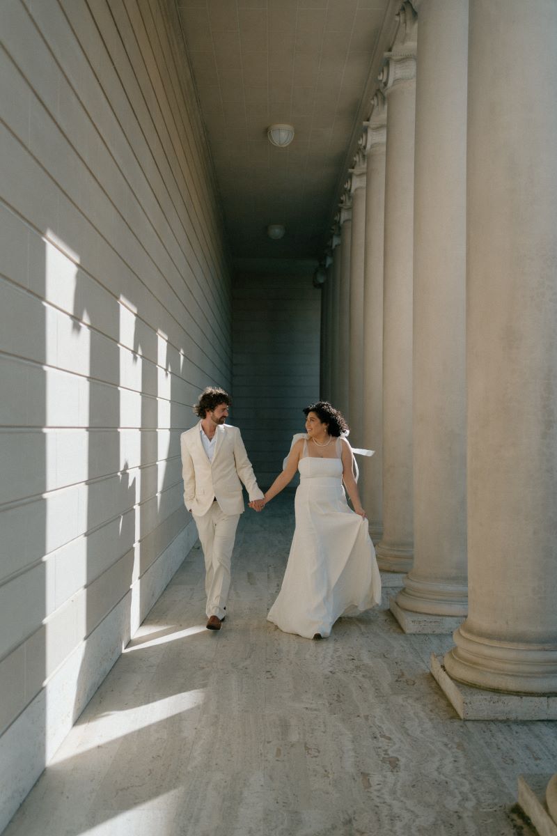 A woman in a white wedding dress is walking hand in hand with her partner who is wearing a tan suit they are both looking at each other and smiling 