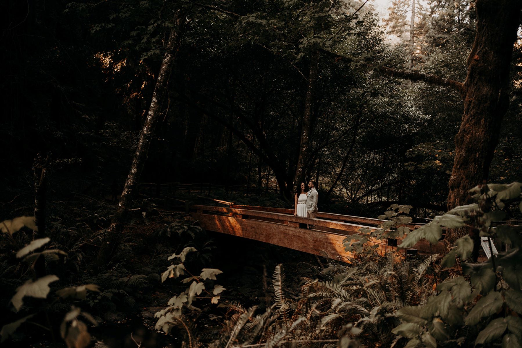 A woman in a white dress in the woods who is looking at her partner who is wearing a jacket and tan pants and they are standing on a wooden bridge surrounded by trees and ferns 