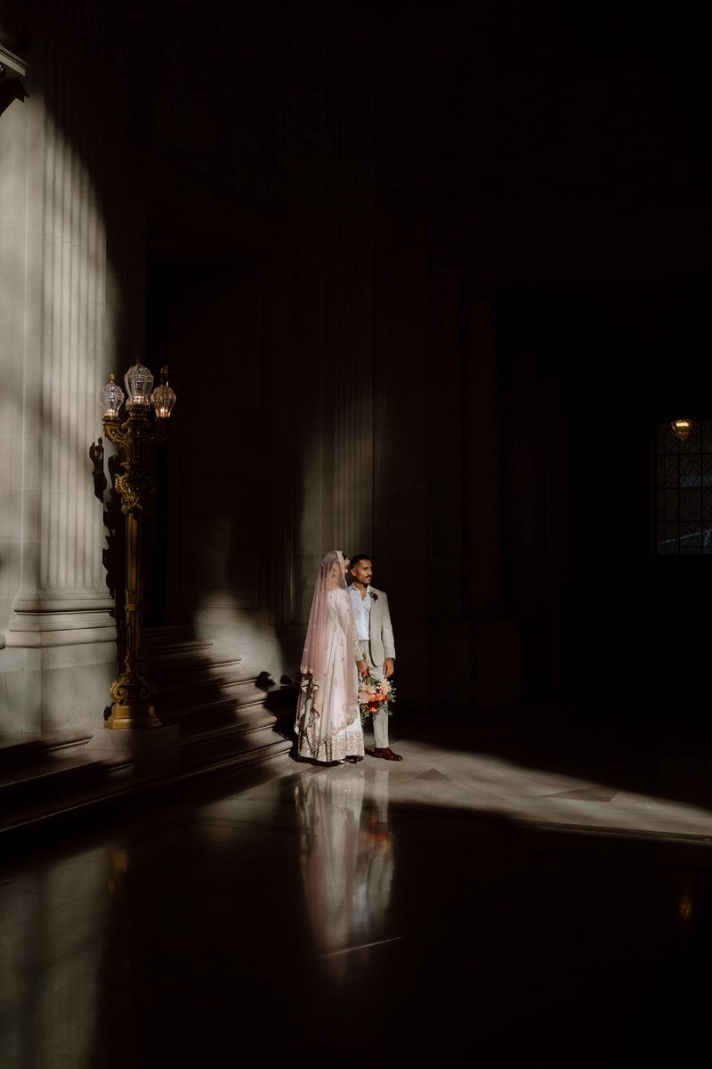 A man and woman in San Francisco City Hall the woman has on a pink veil and a white embroided dress and is holding flowers in her hand and the man is wearing a tan suit and a white button up 