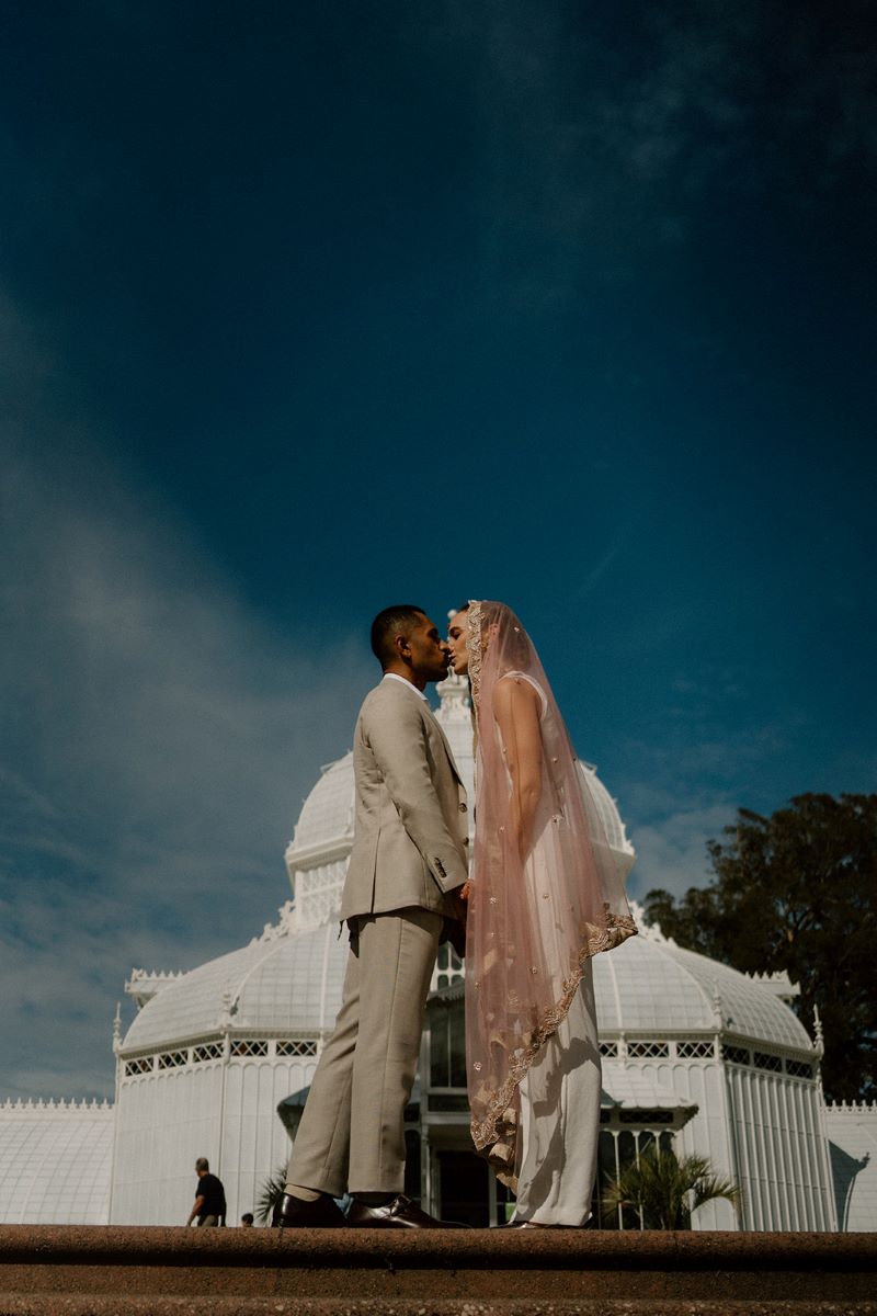 A woman in a pink headress and a white full body suit is giving her partner a kiss at the top of a set of stairs and her partner is wearing a tan suit and behind them is a white conservatory