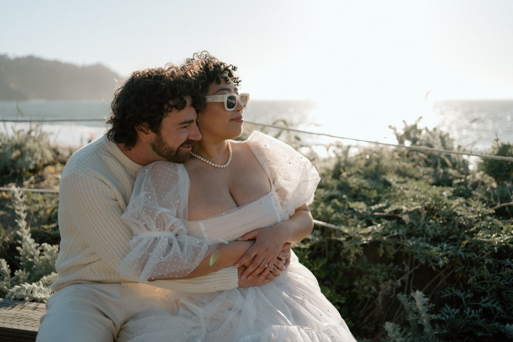 A woman in a white dress and sunglasses and who is wearing a pearl necklace is sitting next to her partner who is wearing a sweater and both are looking off to the distance and behind them is the ocean 