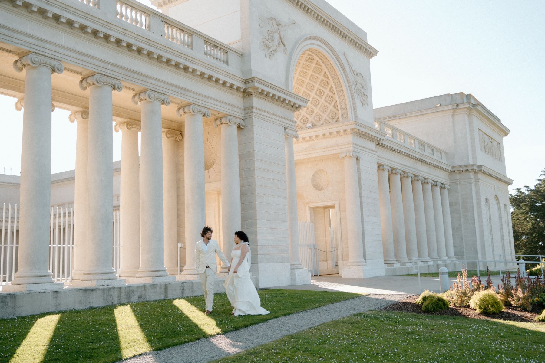 A woman in a white wedding dress walking and holding hands with her partner who is wearing a tan suit and behind them is the Palace of Fine Arts in San Francisco 