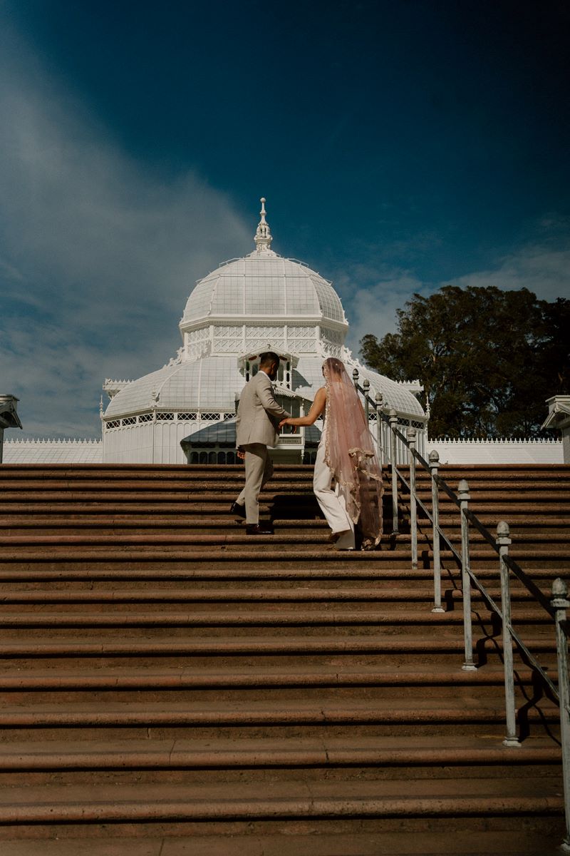 A woman wearing a pink headress and a white body suit is walking up a set of stairs with her partner who is wearing a body suit and behind them is a white conservatory 