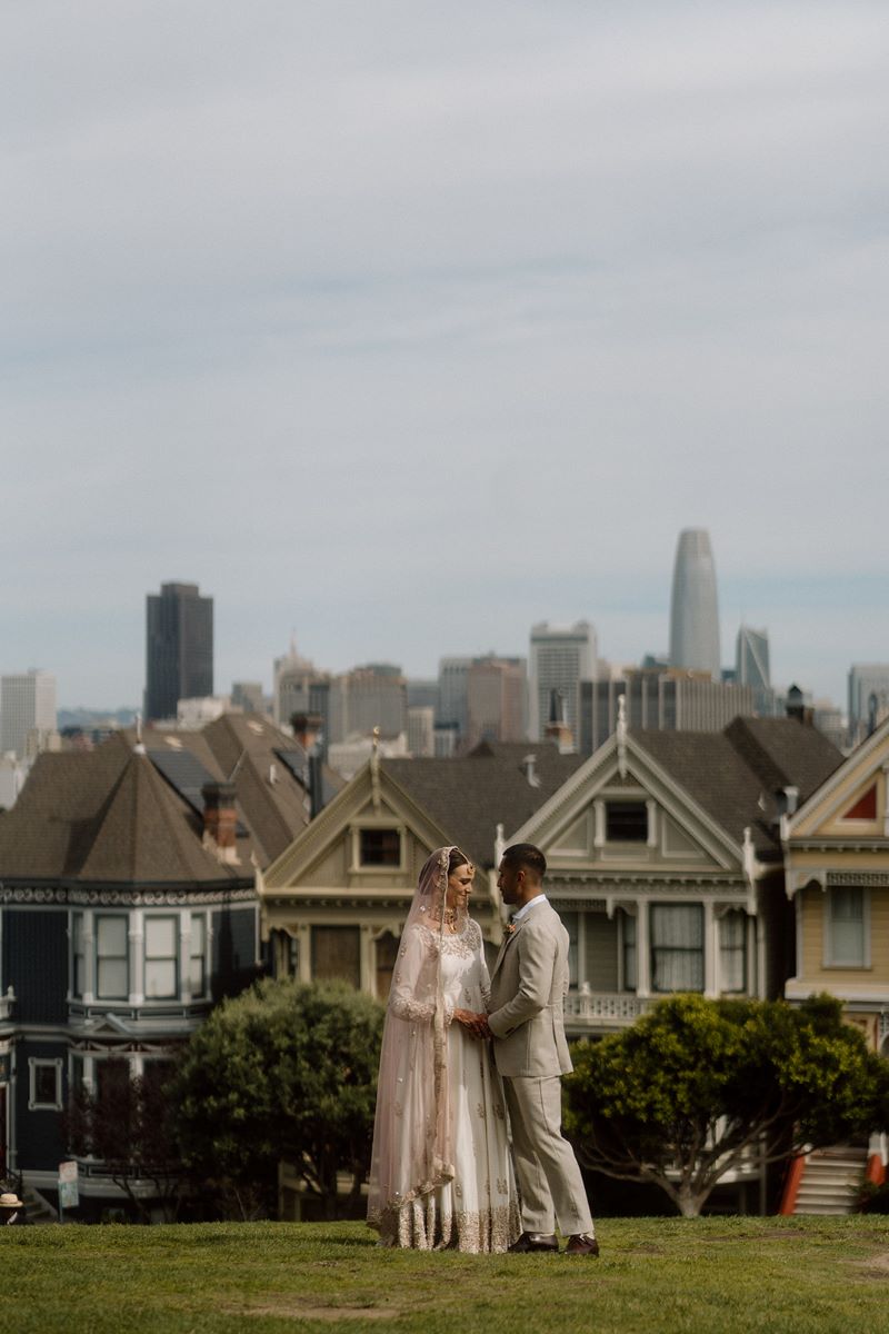 A woman in a pink headress and white dress is holding hands with her partner who is wearing a tan suit and they are both looking at each other behind them are houses and the city of San Francisco 