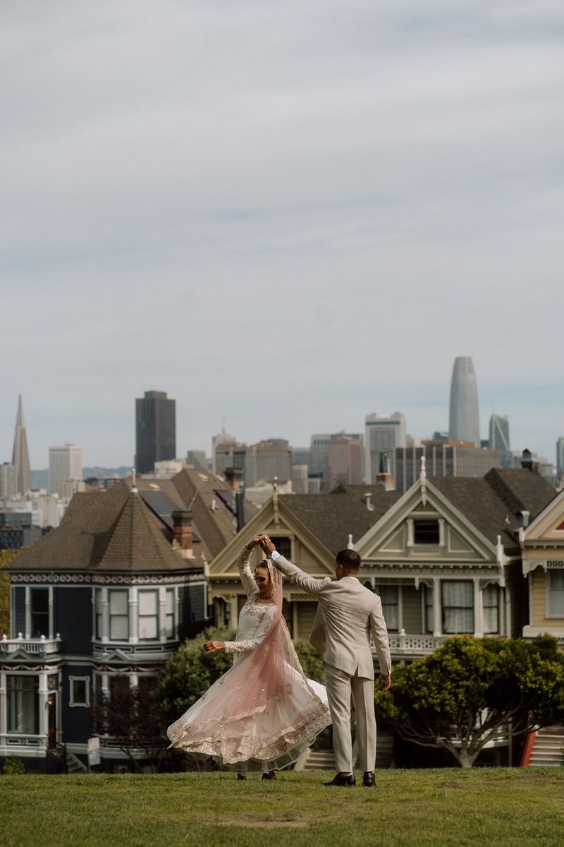 A man in a tan suit is twirling his partner who is wearing a white dress and a pink headress and behind them is the city of San Francisco and some houses