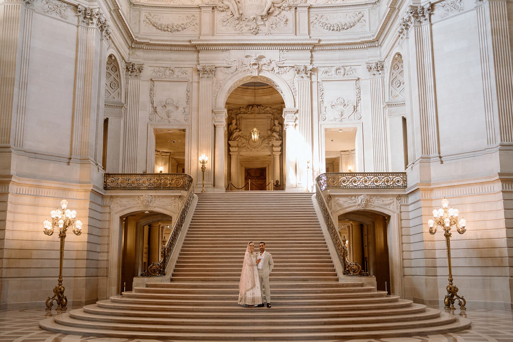 couple at San Francisco City Hall standing on the grand staircase the woman is wearing a wedding dress and a pink veil and the man is wearing a tan suit with a white button up underneath 