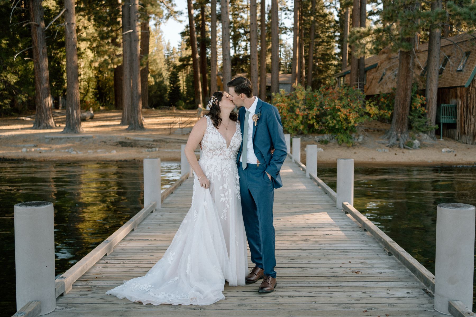 a bride in a white wedding dress giving her husband in a blue suit a kiss they are standing on a dock surrounded by water and trees 