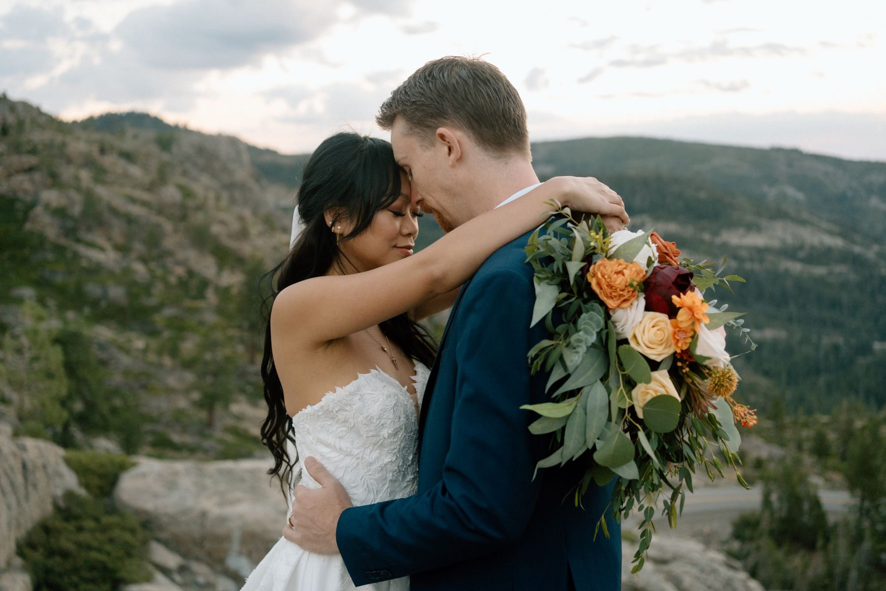 A woman in a white wedding dress pressing her forehead against her partner's forehead who is wearing a blue suit the woman has her arms wrapped around his shoulders and is holding a boquet of flowers with the mountains in the background 