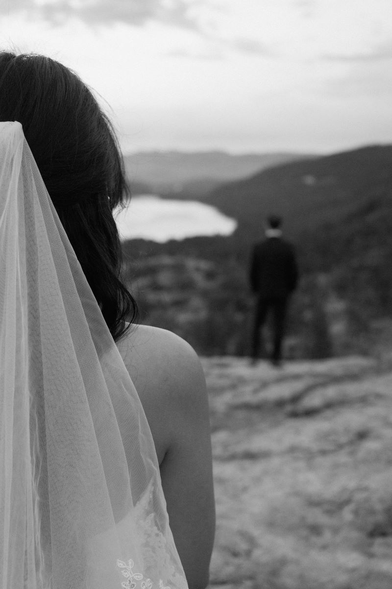 A woman in a wedding dress with a veil looking at her partner who has his back turned and is looking at the mountains 