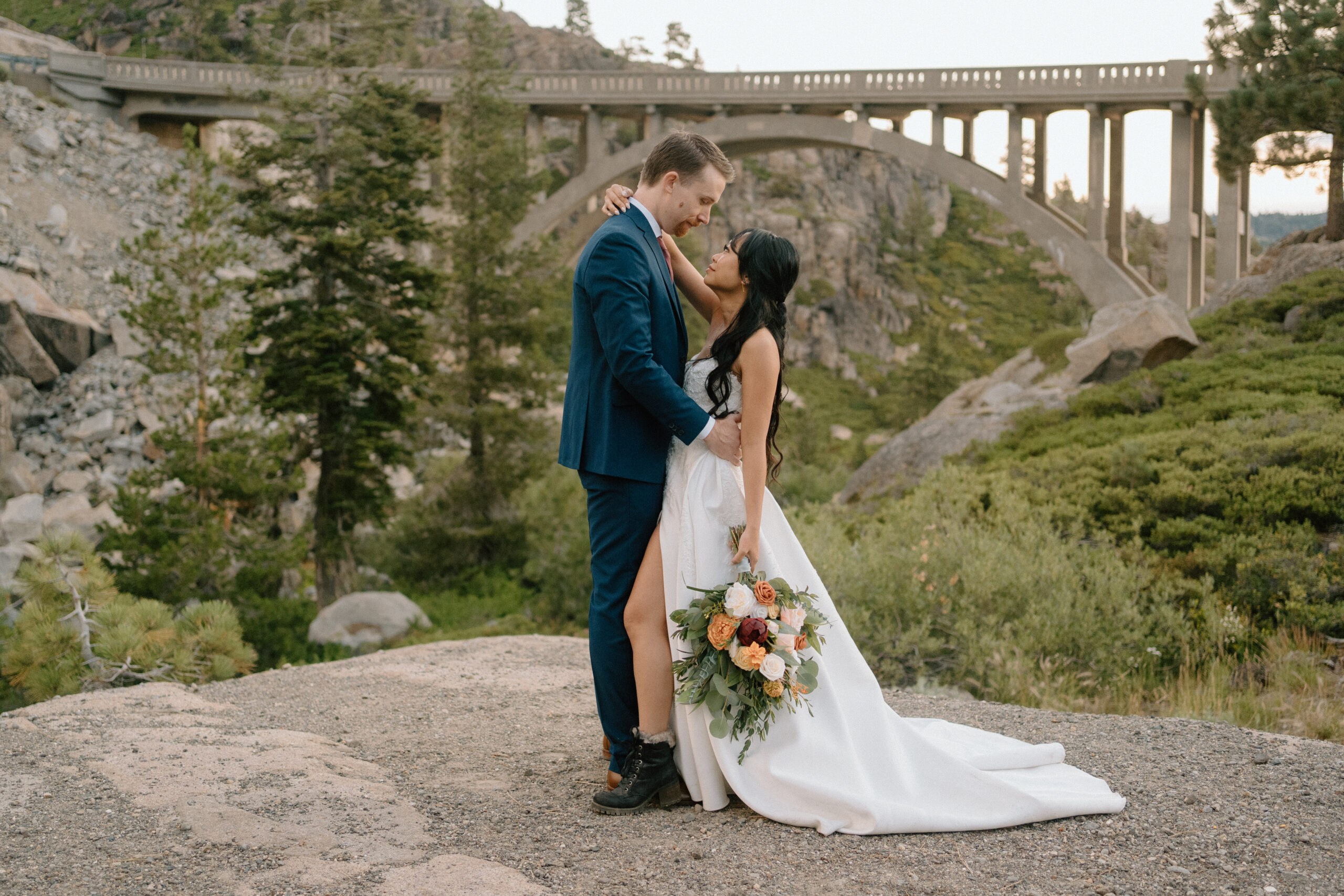 woman in a white wedding dress holding a boquet of flowers in her hand and an arm around her partner's shoulder she is looking up at her partner who is wearing a blue sit and in the background is a bridge