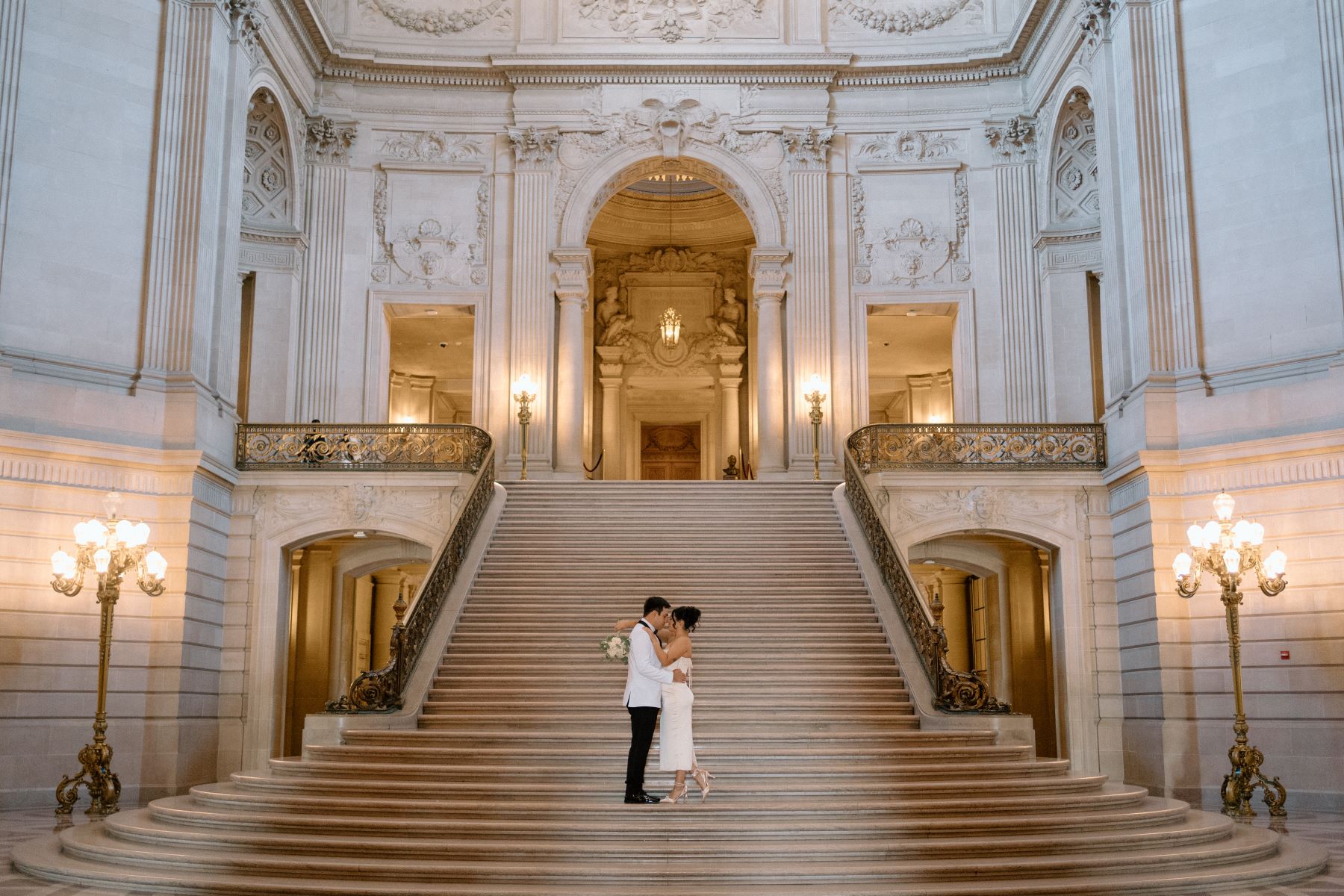 A woman in a white dress and heels standing at the base of San Francisco city hall with her partner who is wearing a white suit 