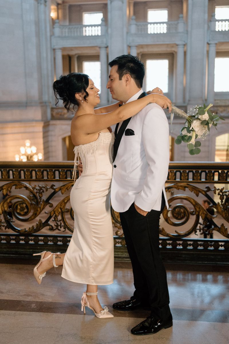 A woman kicking her heel she is wearing a white dress and has her arms wrapped around her partner's shoulder her partner is wearing a white blazer and black pants 