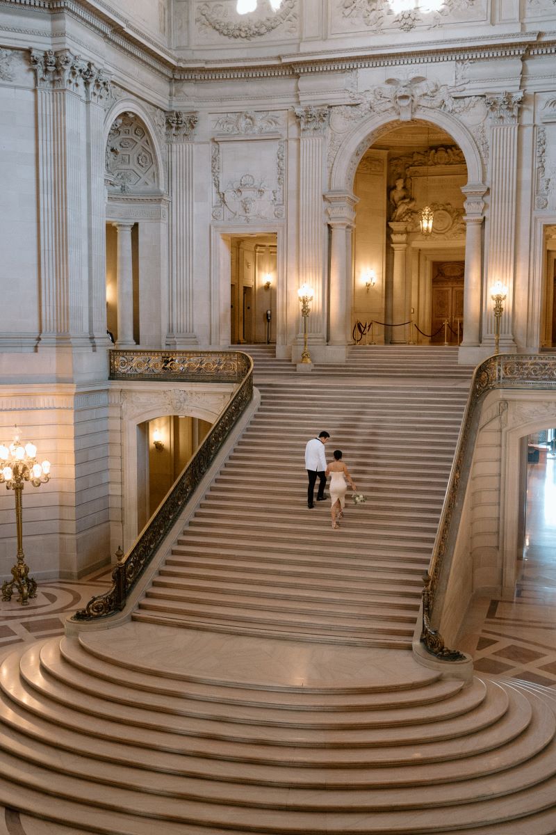 A couple walking up San Francisco city hall's steps the woman is in a dress and the man is wear a white blazer and black pants 