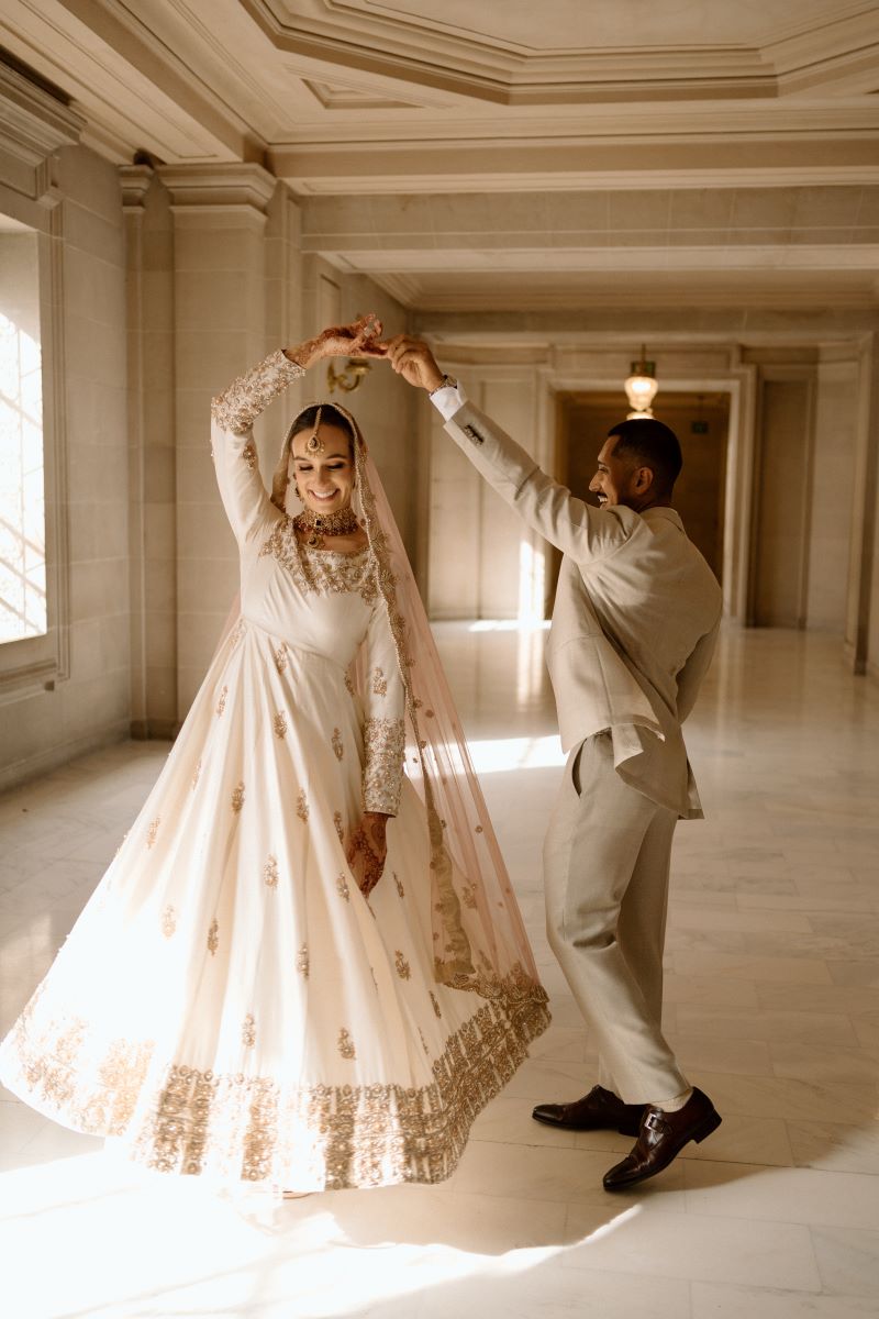 A man in a tan suit twirling in a circle a woman in a white dress with gold embroidery 