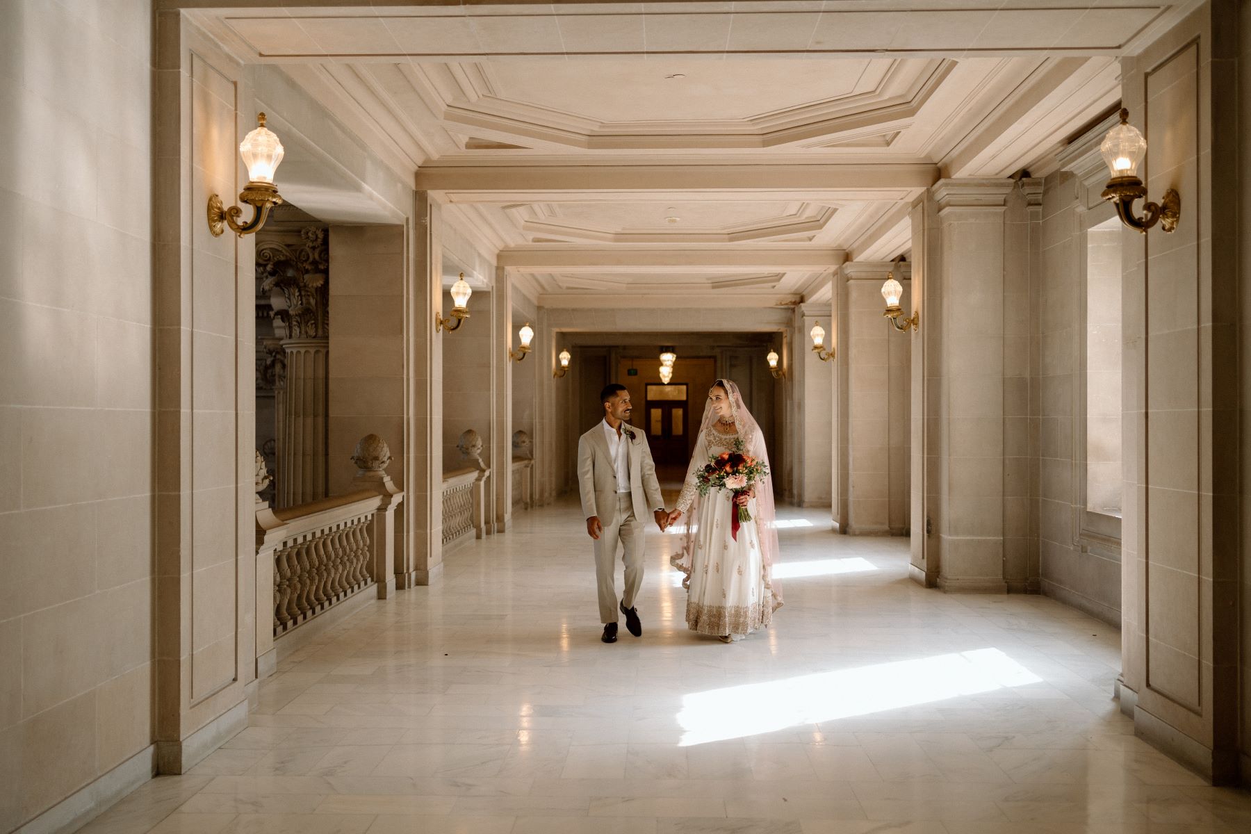 A woman in a white dress with gold embroidery and a pink headress holding a boquet of flowers and looking at her partner who is wearing a tan suit they are walking down the hall and holding hands 