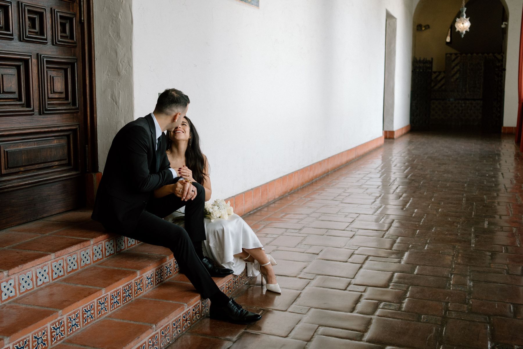 A couple sitting on steps together looking at each other the woman is wearing a white wedding dress and white heels and has a bouquet of flowers in her lap and the man is wearing a black suit