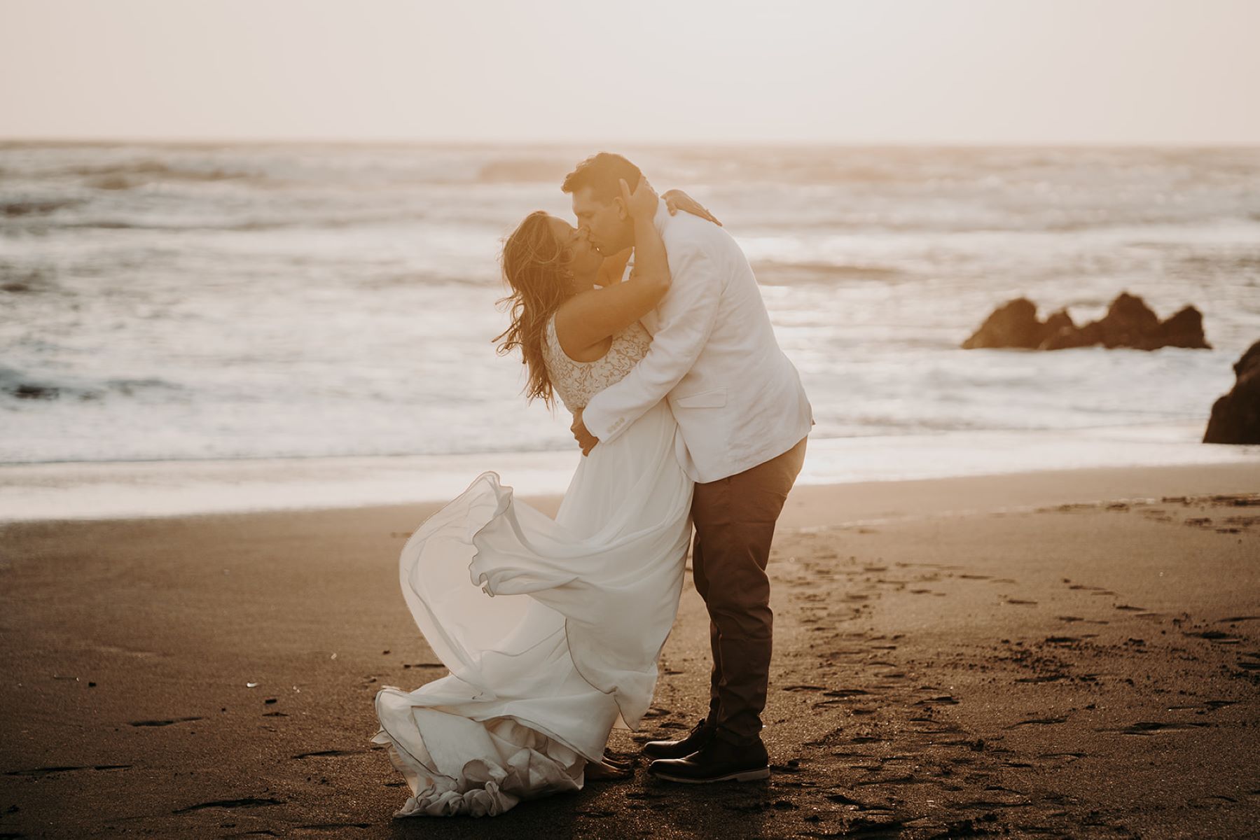 A man in a white jacket and brown pants has his arms wrapped around the waist of a woman wearing a white wedding dress the woman is touching the back of the man's shoulder with one hand and with the other is touching his head and they are both kissing each other on the sand at sunset with the ocean behind them 