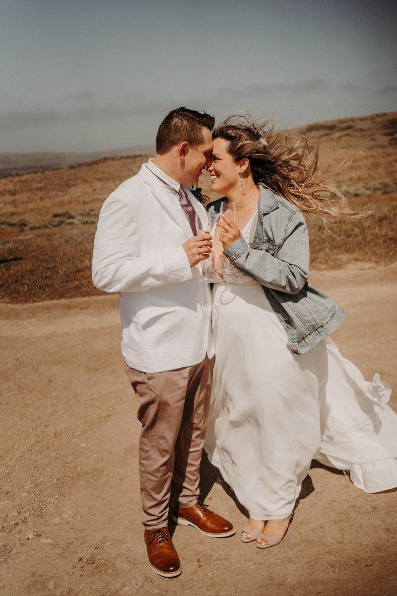 A woman in a jean jacket wearing a white dress is holding a champange glass and touching her head to her partner's who is wearing a white suit jacket and brown pants and is also holding a champange glass 