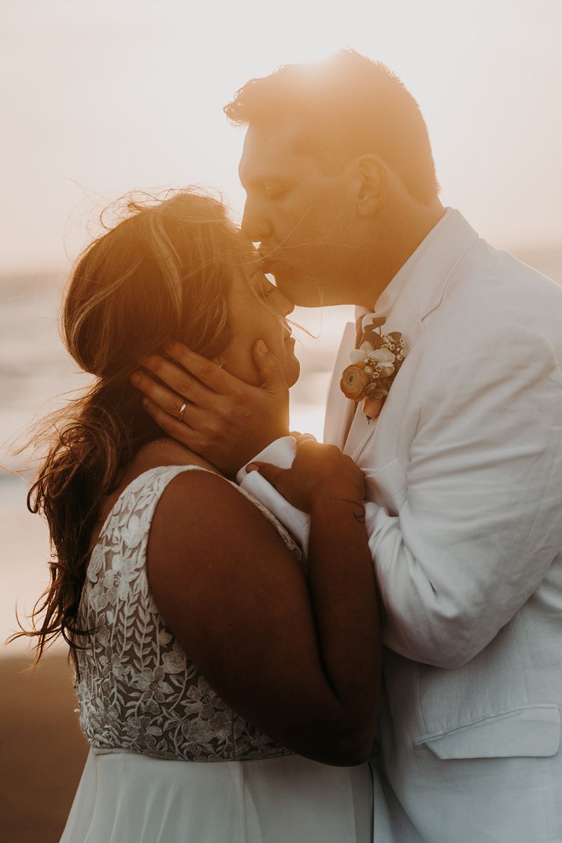 A woman in a white wedding dress is holding the wrists of a man wearing a white suit jacket who is kisisng her forehead as the sun sets and the ocean is behind them 