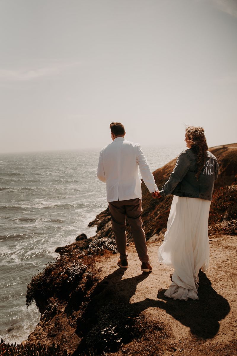 A woman in a jean jacket and white wedding dress is holding hands with a man wearing a white suit jacket and brown pants they are are both looking out toward the ocean and standing on a cliff 