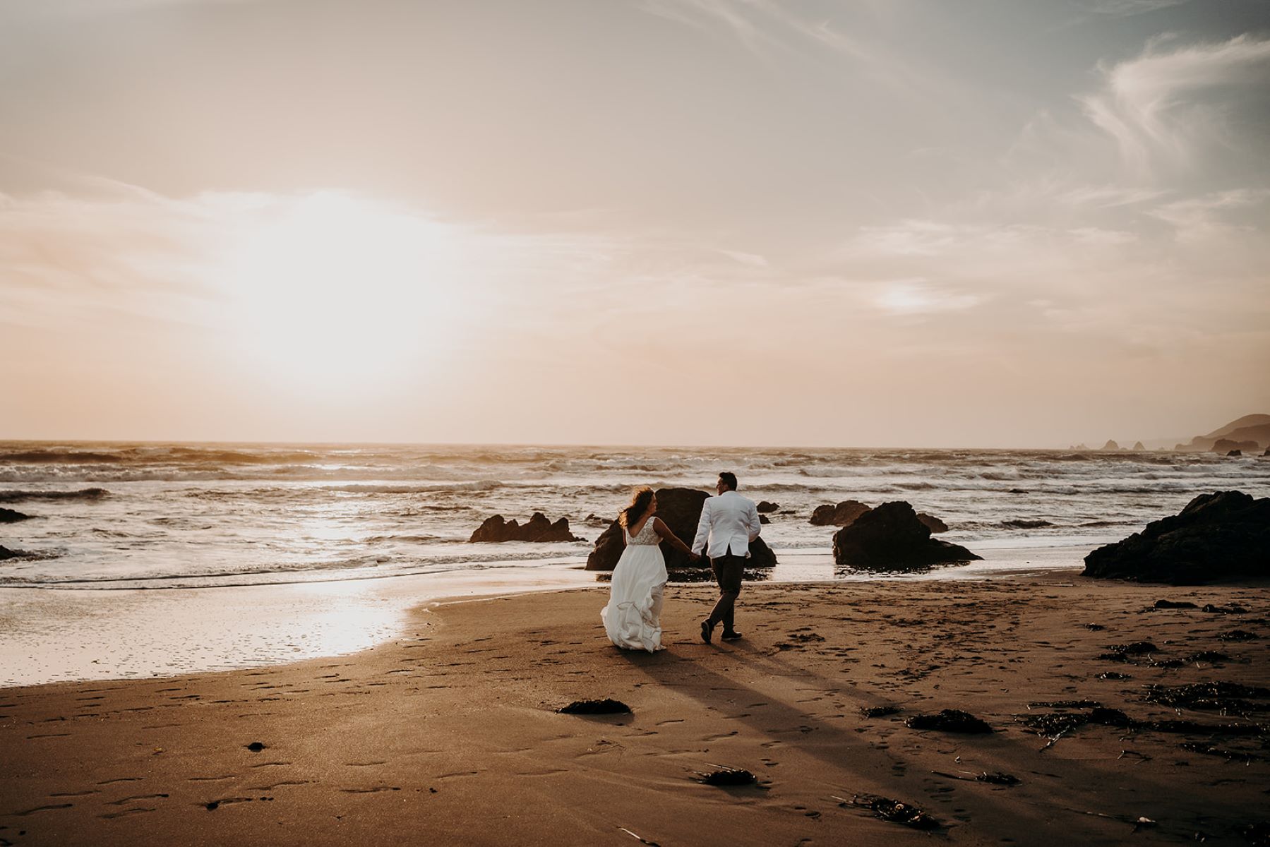 A man in a white suit jacket and brown pants is running on the beach and holding hands with his partner who is wearing a white wedding dress next to the ocean and some rocks