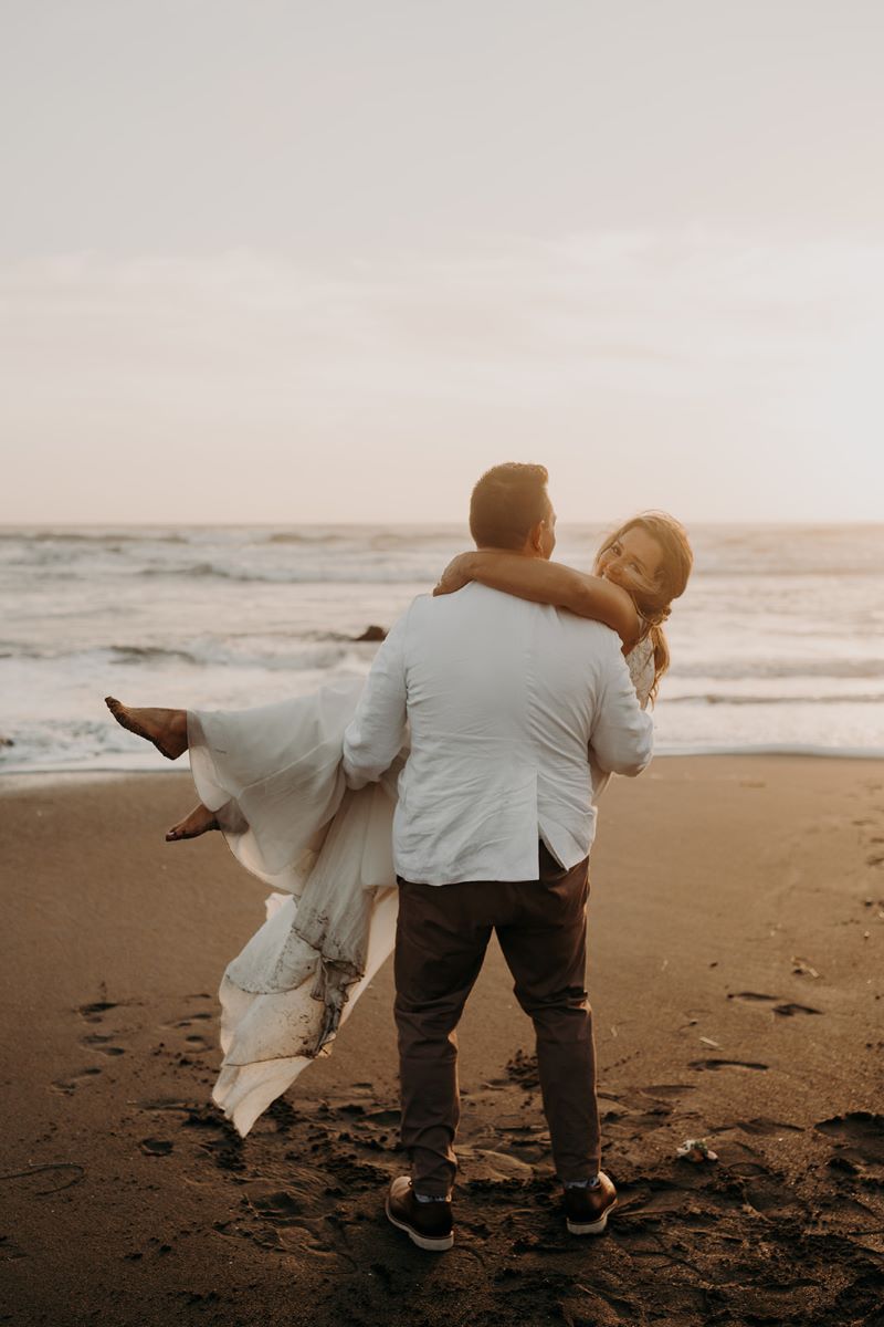 A man in a white suit jacket and bronw pants is standing on the sand next to the ocean and is lifting his wife in his arms who is wearing a white wedding dress and smiling 