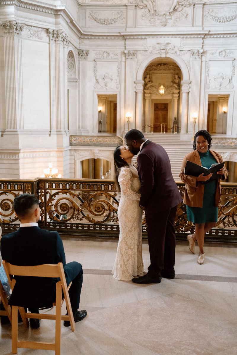 A couple kissing at their wedding ceremony their officiant is smiling in the background and their guests are watching them the woman has on a white dress and the man is wearing a maroon tuxedo