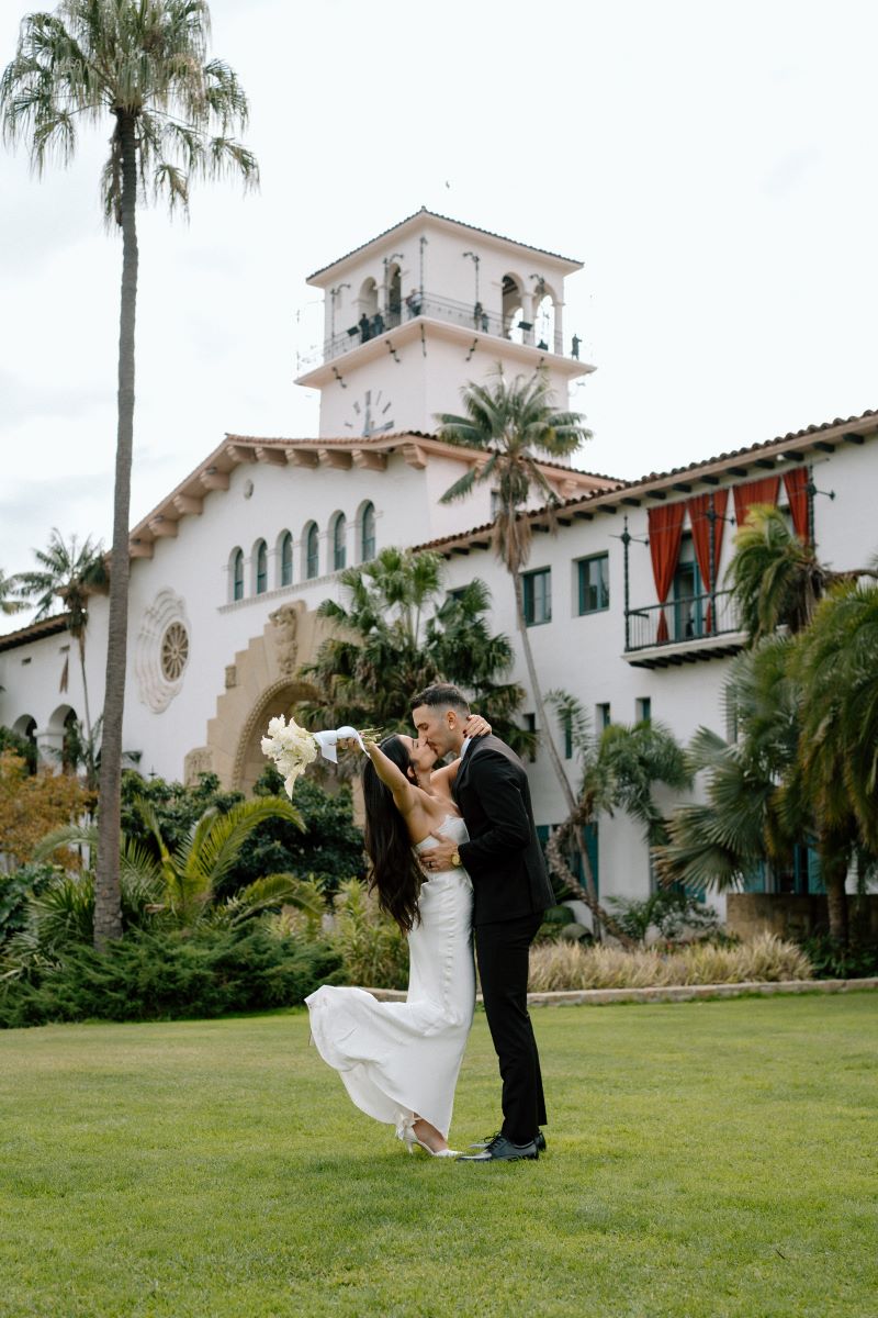 A woman in a white wedding dress and heels is kissing her partner and kicking up her heel in the air and is holding her arm up while holding a bouquet of flowers and her partner is wearing a black suit and kissing her behind them is a white hacienda style house 