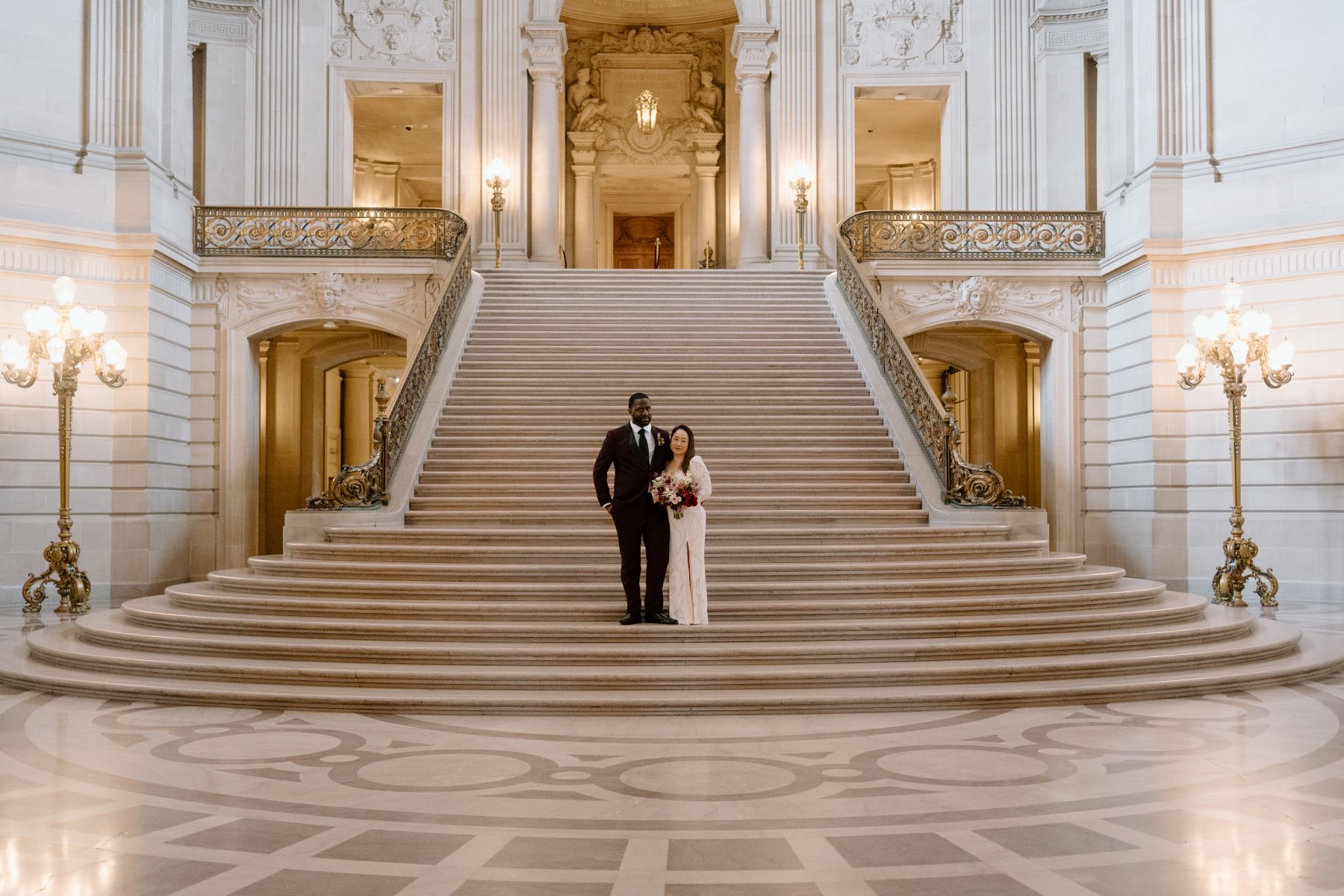 A couple standing ona grand staircase the woman is wearing a white wedding dress and holding a bouquet of flowers and the man is wearing a maroon tuxedo and has his hand on his partner's back 