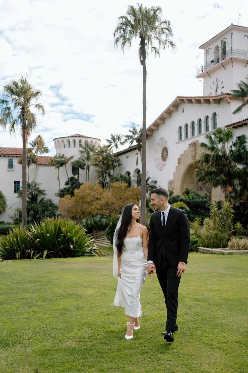 A woman in a white wedding dress with a white veil and white heels is holding hands with here partner and looking at him and smiling and her partner is wearing a black suit and looking at her and smiling behind them is a white hacienda style house and lots plants 