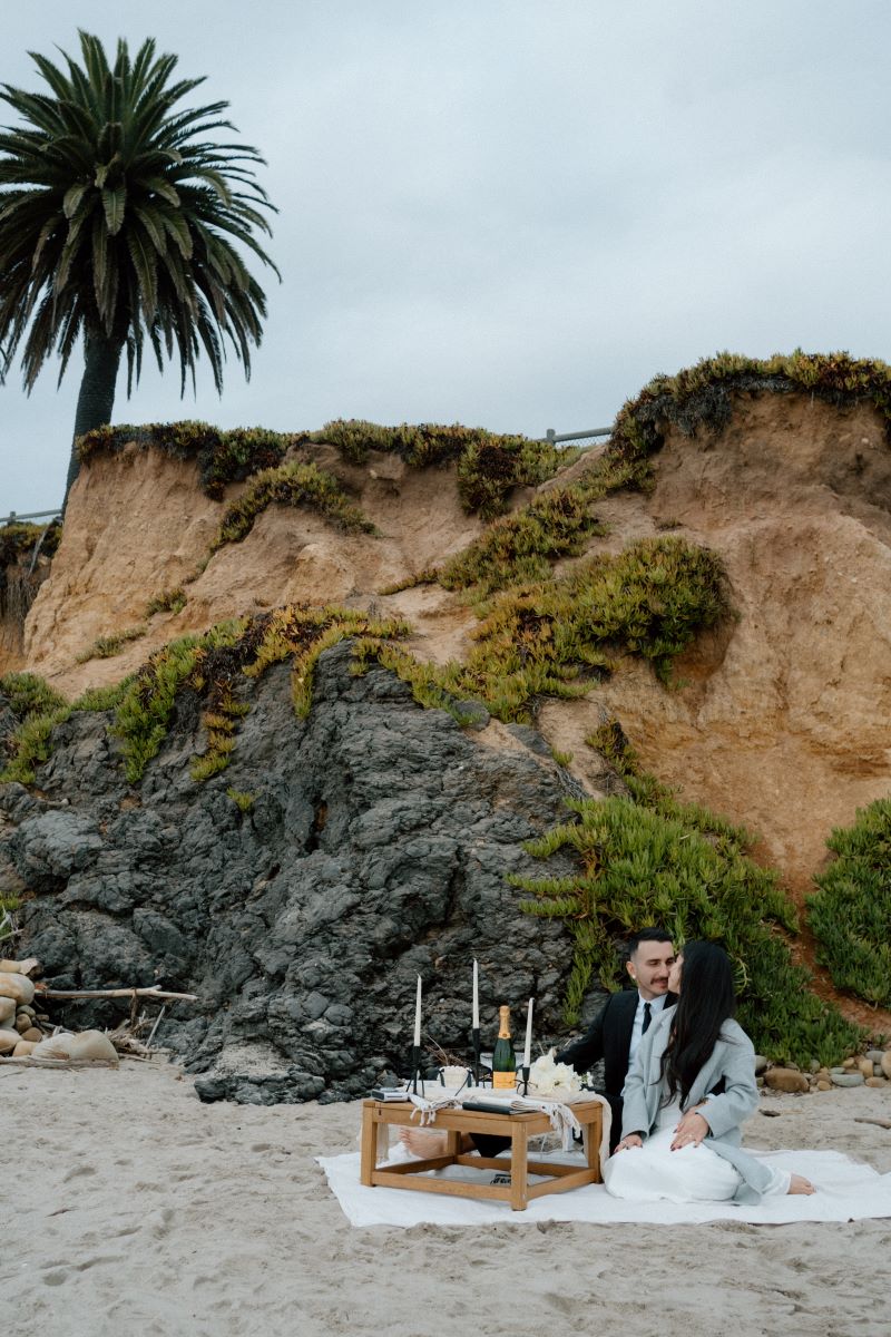 A couple enjoying a luxury picnic at the beach sitting on a white blanket and looking at each other in front of them is a table with candles and food and bheind them are rock formations 