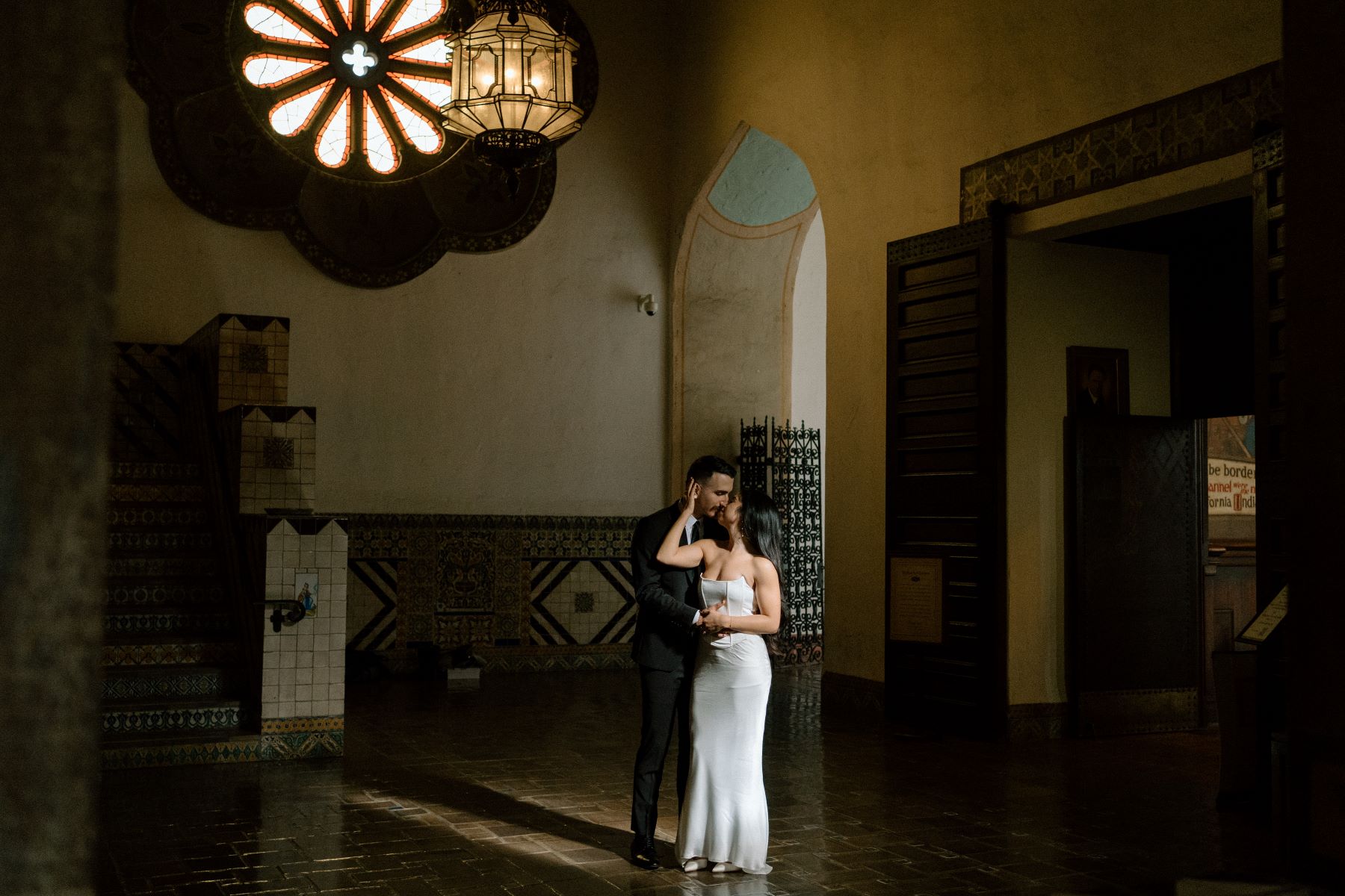 A couple looking at each other in a dark hall the woman is wearing a white wedding dress and is touching her partner's face with one hand and her partner is wearing a black suit and holding her hand