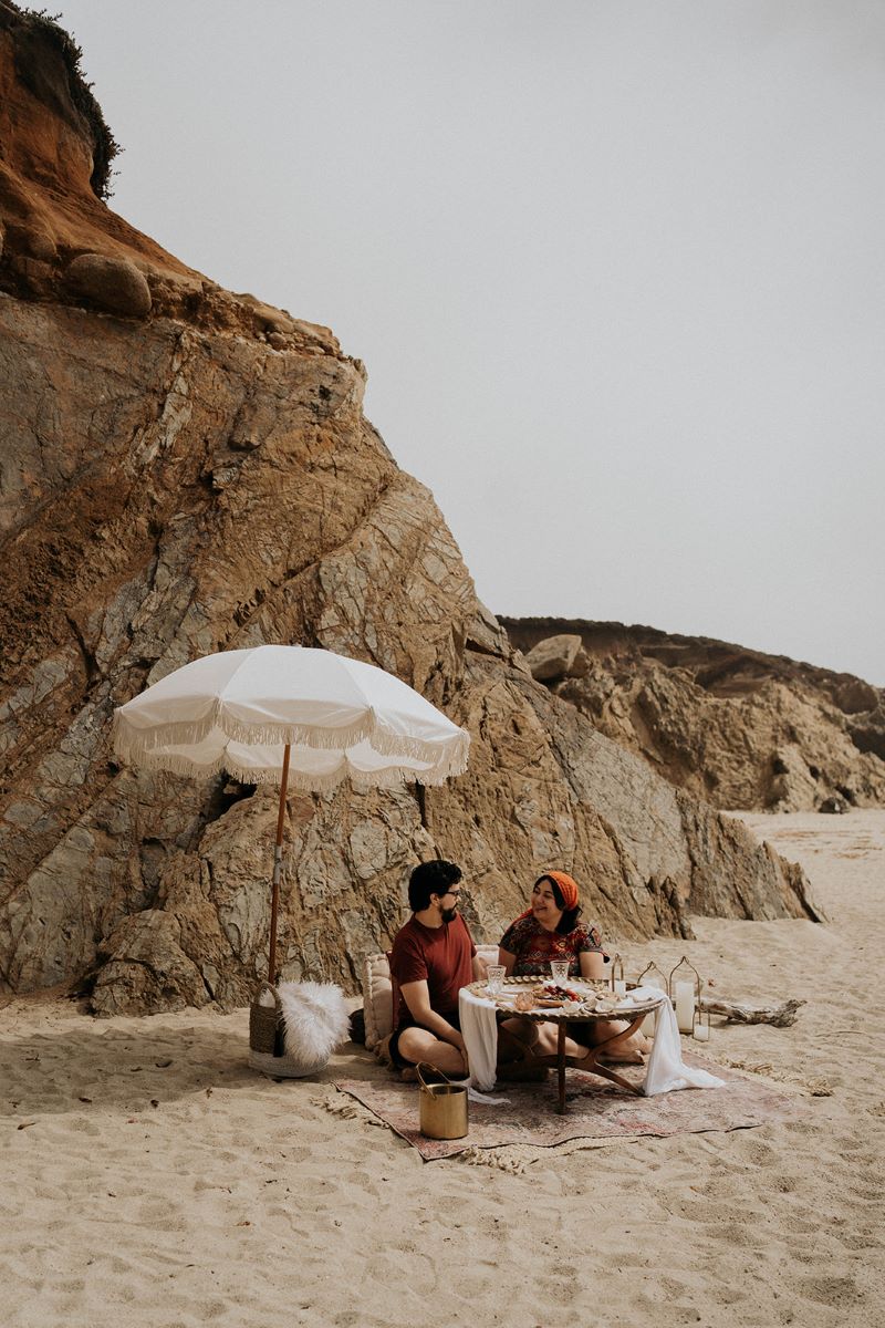 A couple enjoying a luxury picnic at the beach in the sand behind them are rock formations and in front of them is a table and behind them is a white umbrella the couple is sitting on a blanket and laughing with each other