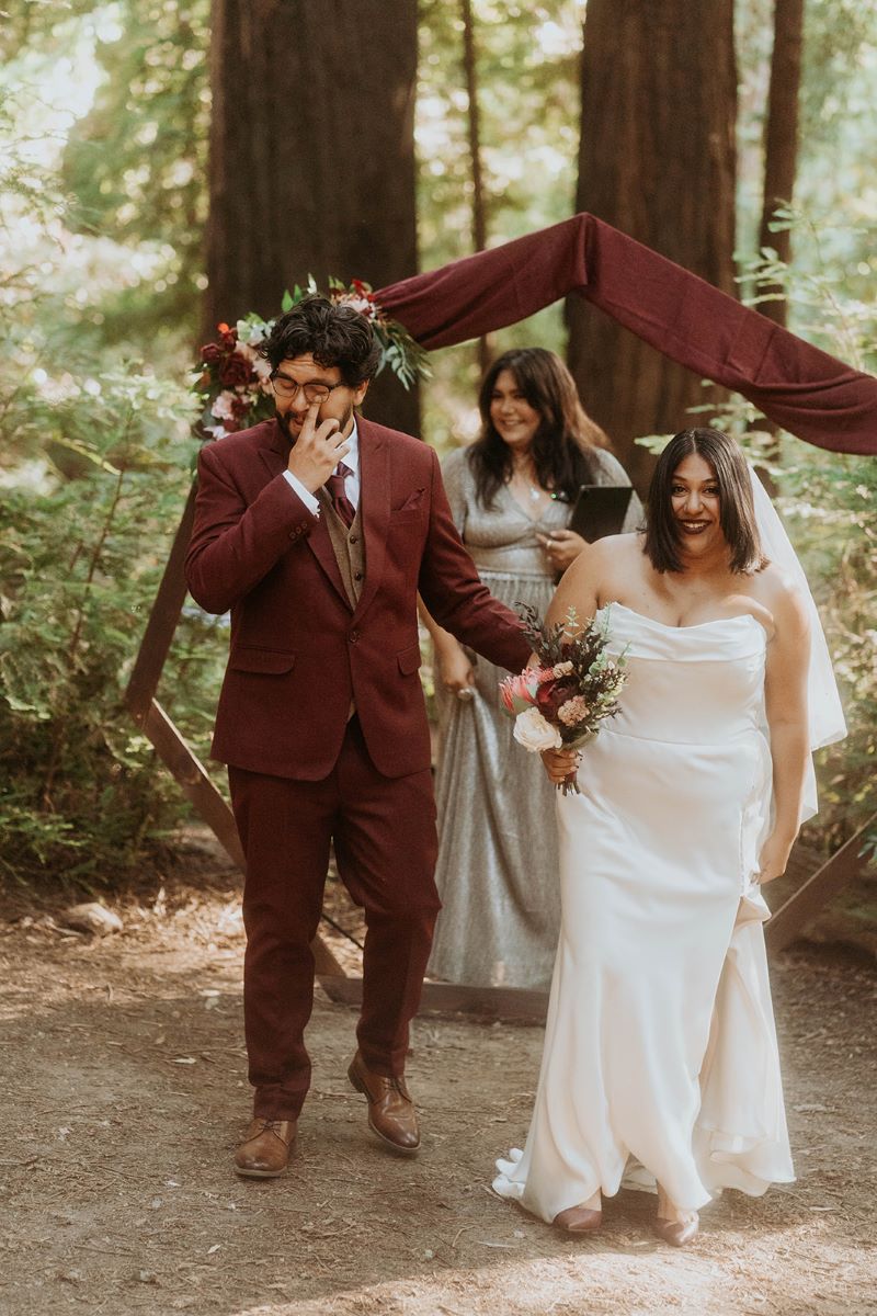 A woman in a white wedding dress with a white wedding veil is holding a bouquet of flowers and her partner is wearing a red suit and has glasses and is touching her back and is wiping away a tear from his eye after their wedding ceremony and their officiant is standing in the background