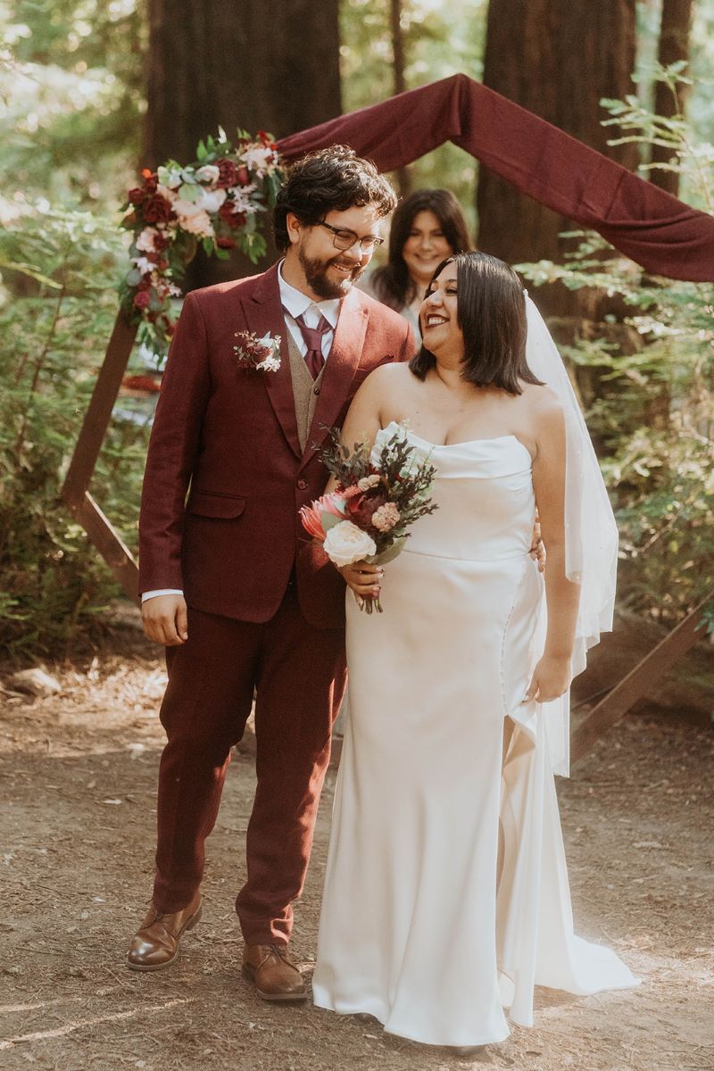 A woman in a white wedding dress and a white veil is holding a bouquet of flowers and smiling and looking at her partner who is wearing a red suit and has glasses and is looking at her they are in front of the ceremony arch and their wedding officiant is in the background smiling they had their wedding ceremony in a forest