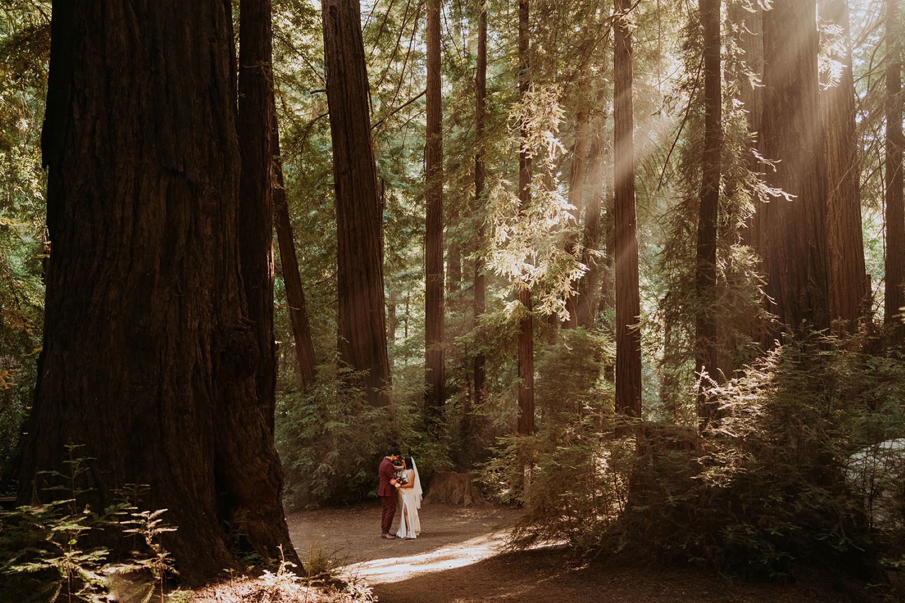 A couple looking at each other in a forest the woman is wearing a white wedding veil and dress and holding a bouquet of flowers her partner is wearing a red suit