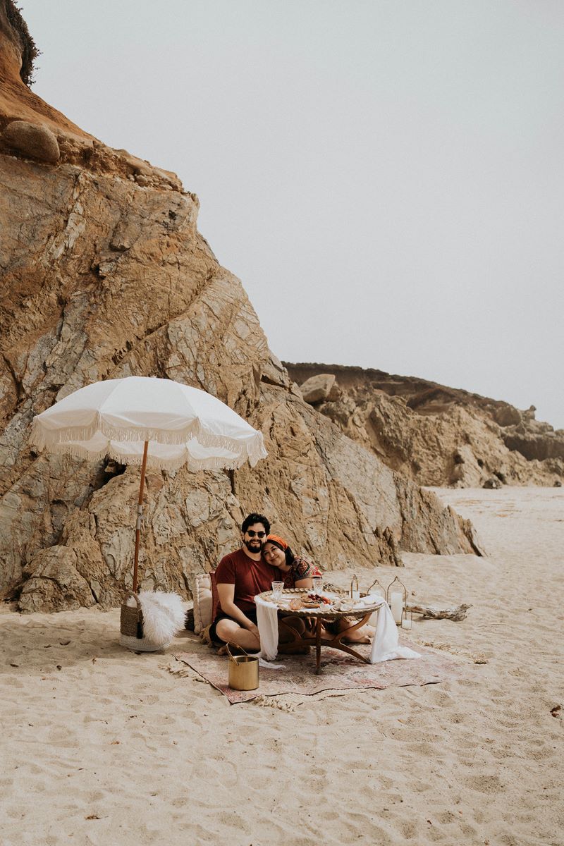 A couple enjoying a luxury picnic at the beach in the sand behind them are rock formations and in front of them is a table and behind them is a white umbrella the couple is sitting on a blanket and the woman has her head on her partner's shoulder 