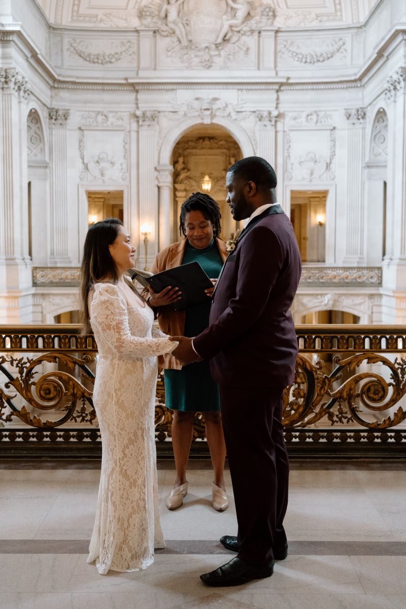 A couple holding hands at their wedding ceremony they are looking at each other and their officiant is behind them the woman has a white wedding dress and the man has on a maroon tuxedo 