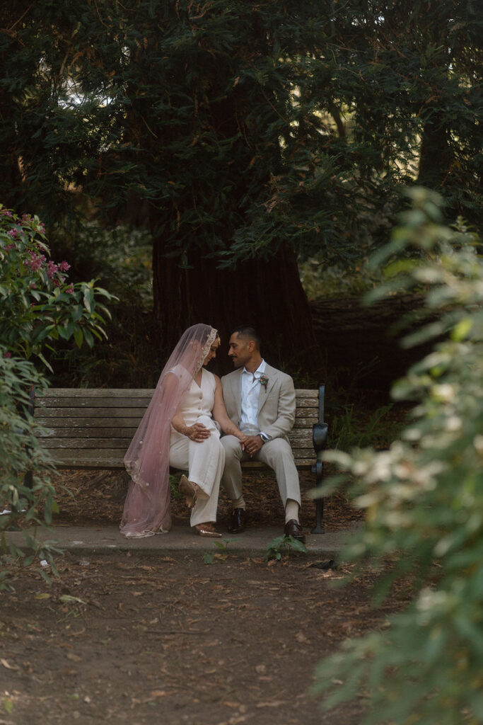 A newlywed couple sitting on a bench in a park looking at each other 
