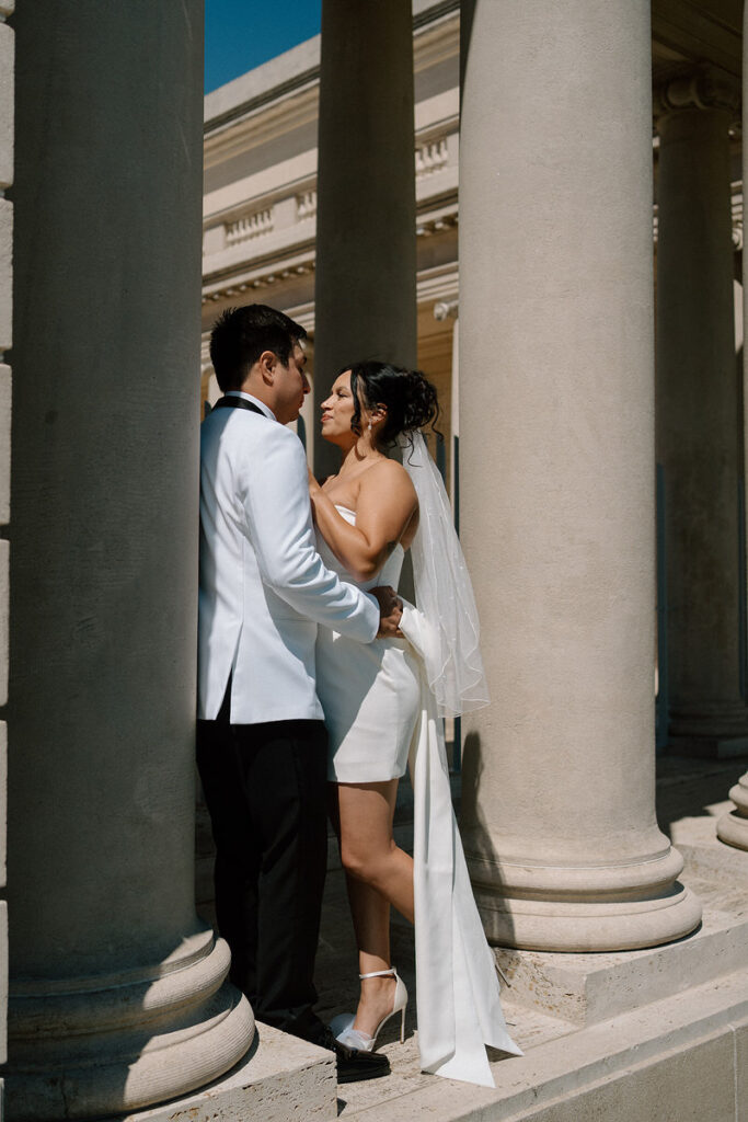 A newlywed couple standing close and leaning on a large pillar to a building 