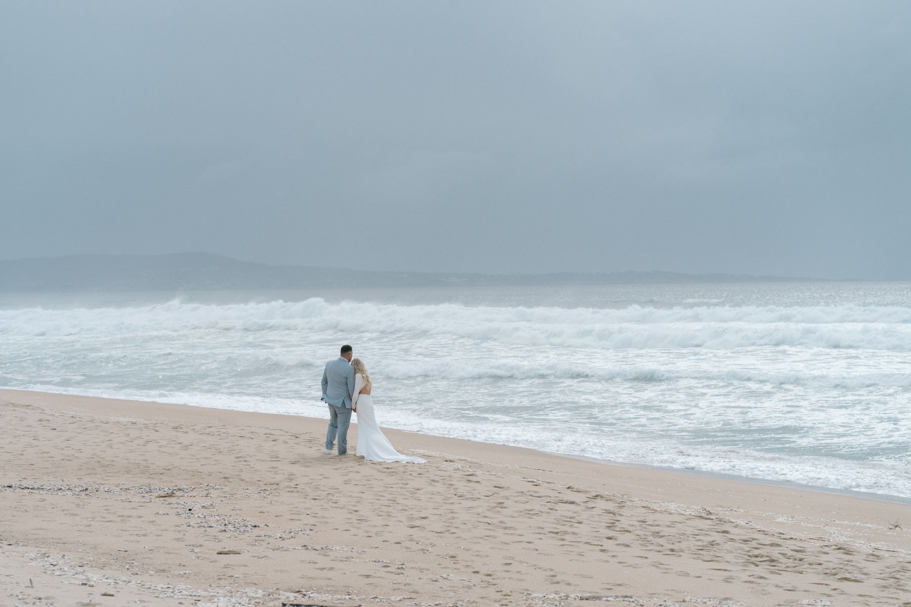 A woman in a white wedding dress and a man ina blue suit holding hands on the sand and looking at the ocean together