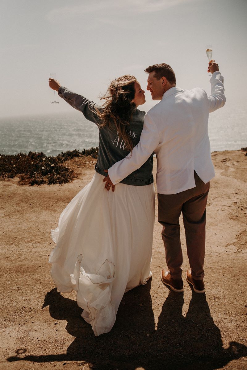 A couple standing in front of the ocean holding champagne glasses in the air and toasting the woman is wearing a white wedding dress and a jean jacket the man is wearing a white dress jacket and tan pants 