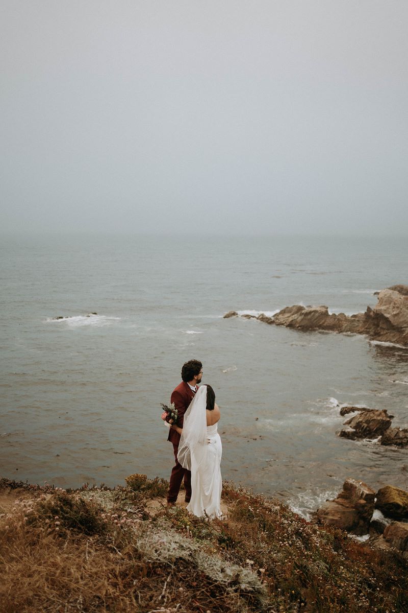 A woman in a white wedding dress with a wedding veil is hugging her partner who is wearing a red suit the woman is also holding a bouquet of flowers they are both standing on a cliff together and looking at the ocean 