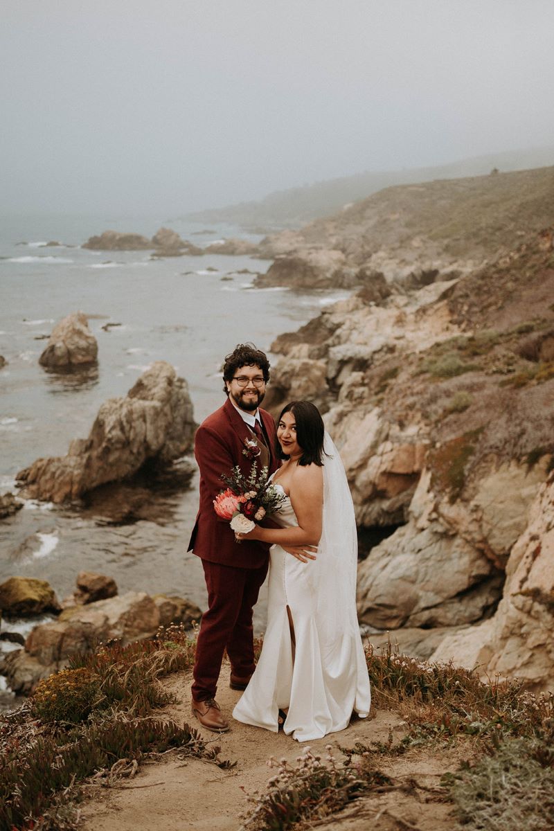 A woman in a white wedding dress with a white veil is holding a bouquet of flowers and standing next to her partner who is wearing a red suit and they are standing on a cliff together and the ocean is behind them 