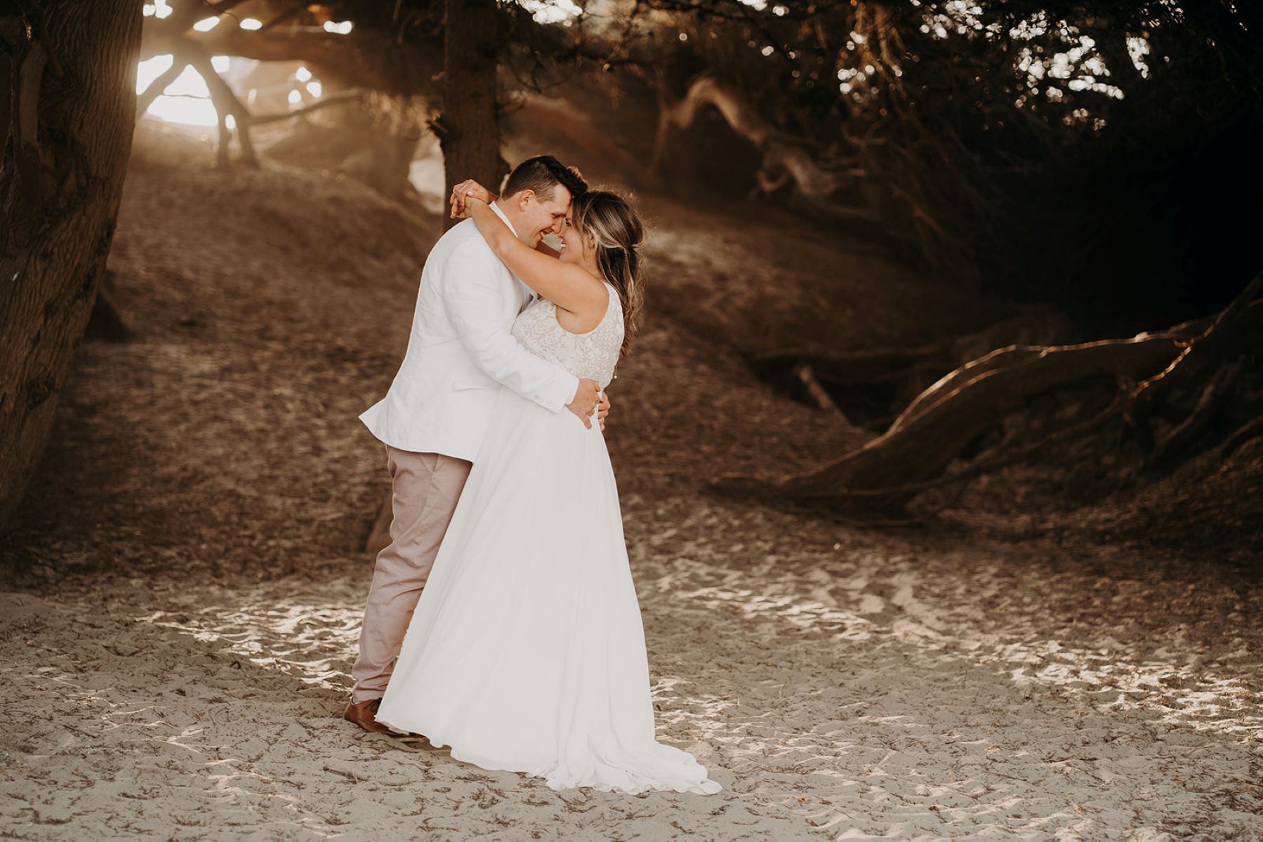 A woman wearing a white wedding dress has her arms around her partner's neck who is wearing a white suit jacket and brown pants and is hugging his partner around her waist they are both standing on sand with a grove of trees behind them