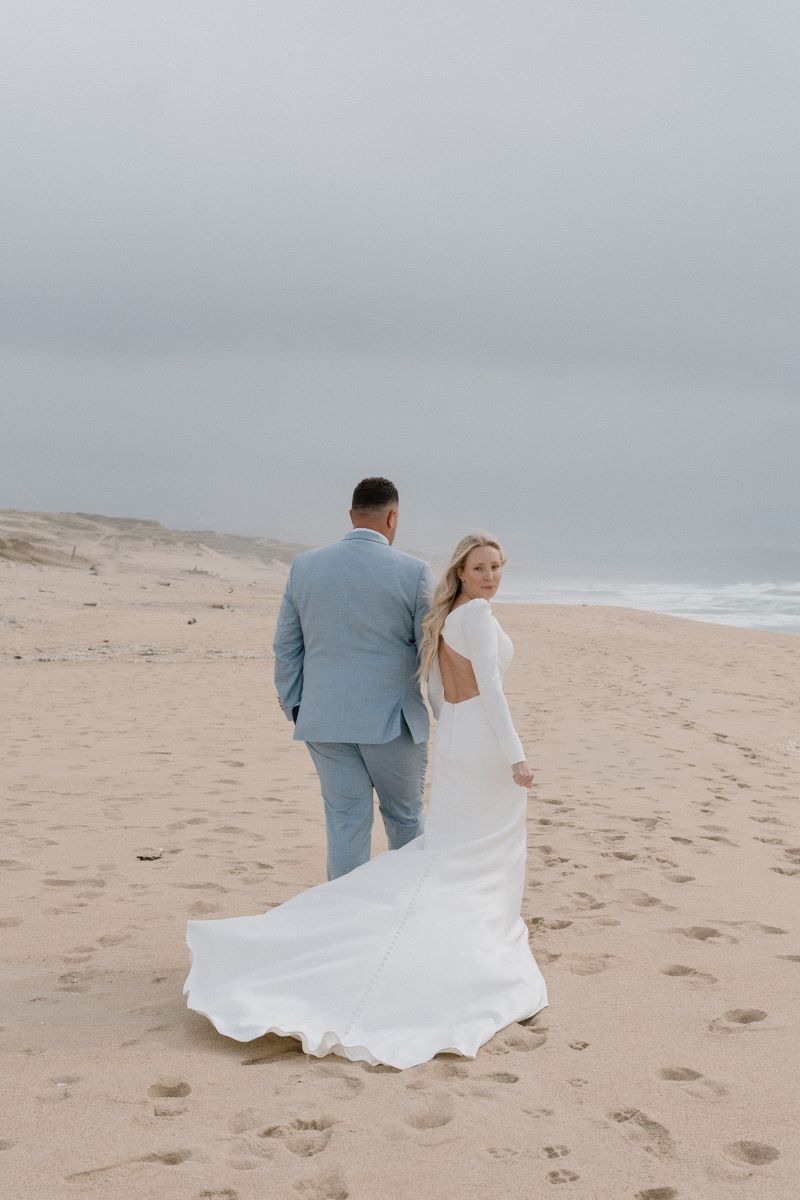 A woman in a white wedding dress is walking on the beach next to the ocean with her partner who is wearing a blue suit